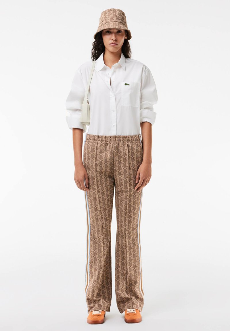 Kobieta w beżowym wzorzystym kapeluszu typu bucket i dopasowanych spodniach, w białej koszuli z guzikami, białej torbie na ramię i pomarańczowych trampkach, stojąca prosto.