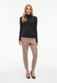 Crna dugorukava majica s visokim ovratnikom, bež uske hlače i crne cipele s visokim potpeticama sa zlatnim detaljima, na jednostavnoj bijeloj pozadini.