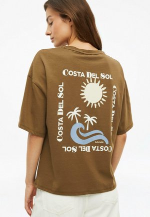 Femme portant un t-shirt marron oversize avec le texte "Costa Del Sol", soleil, palmiers et graphisme de vague dans le dos.