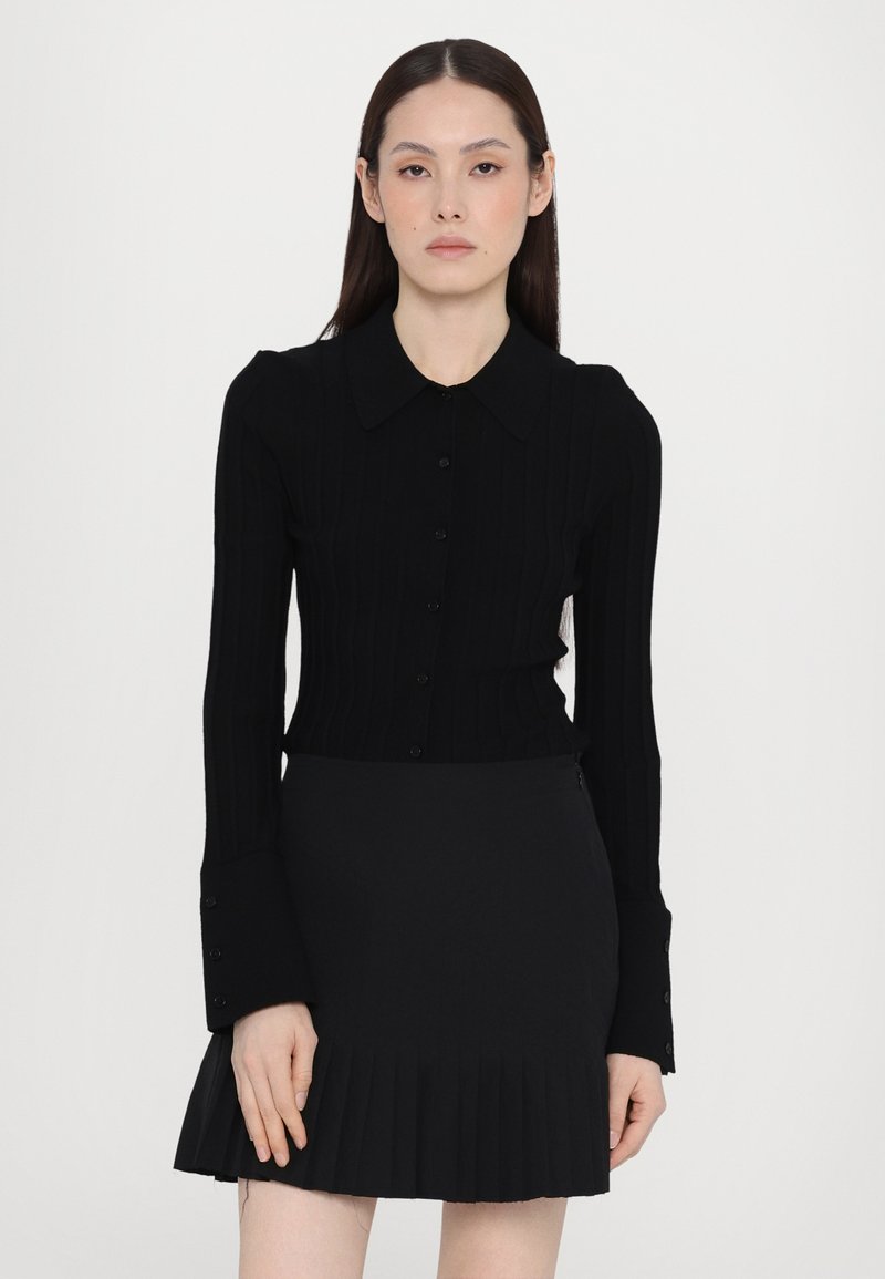 Maglia nera a costine con maniche lunghe, colletto e chiusura a bottoni, abbinata a una gonna plissettata nera. Entrambi gli indumenti in tessuto completamente nero.