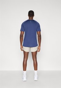 Μαρινί μπλουζάκι γυμναστικής με κοντά μανίκια και διακριτές ραφές, συνδυασμένο με ανοιχτό μπεζ σορτς και λευκά αθλητικά παπούτσια.