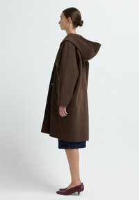 Brauner Wollmischungs-Mantel mit Kapuze, der eine gerade Silhouette, große Ärmel und minimalistische Verschlussdetails aufweist. Kombiniert mit einem Jeansrock und burgunderfarbenen Schuhen.