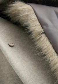 Beige wol stof met een zachte nepbont rand langs de rand, voorzien van een ronde knoopsluiting in een lichte taupe kleur. Glad interieur voering.