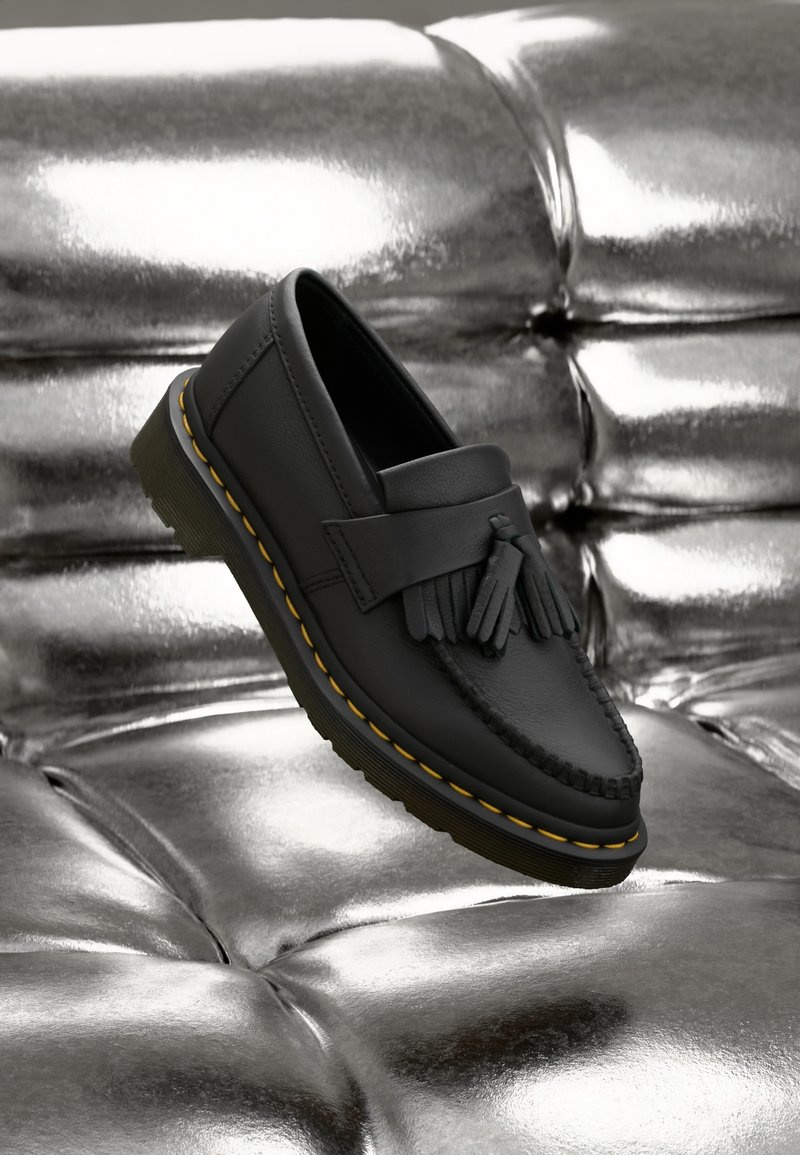 Mocassin en cuir noir avec pompons et semelle jaune cousue, reposant sur une surface argentée tuftée brillante.