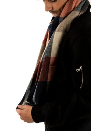 Joven con chaqueta negra y sosteniendo una bufanda a rayas multicolor, mirando hacia abajo contra un fondo blanco.