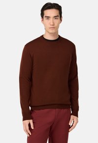 Rjav pleten sveter z okroglim izrezom in dolgimi rokavi, z gladko teksturo in rebrastimi manšetami, v kombinaciji s svojim maroon hlačami.