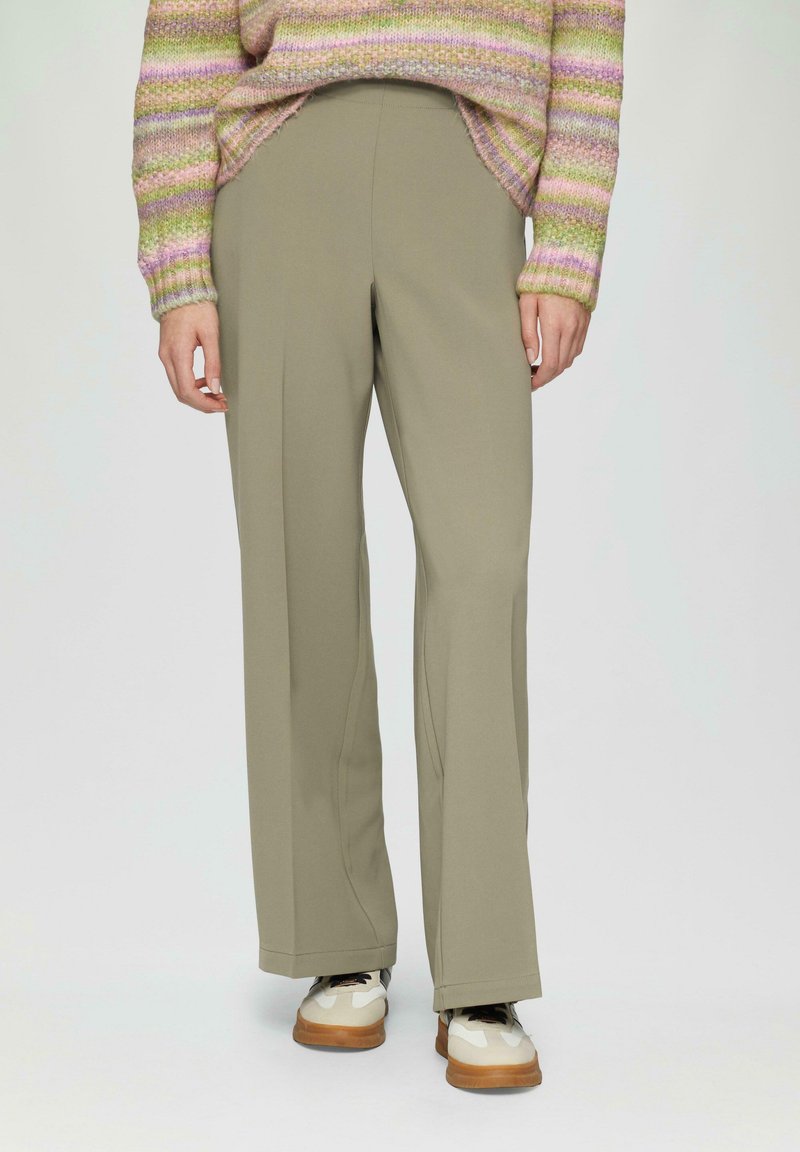 Pantalons à jambe large en vert olive, confectionnés en tissu lisse, avec une coupe ajustée et des coutures latérales. Portés avec des baskets blanches et beige.