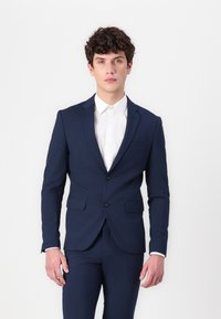 Marineblauer Anzug mit einem taillierten Blazer, Ein-Knopf-Verschluss, zwei Fronttaschen und passenden Hosen. Darunter ein weißes Hemd.