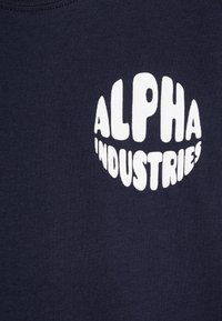 Tamsi mėlyna medvilninė marškinėliai su dideliu baltu apvaliu logotipu, kuriame aiškiai išspausdinta tekstas „ALPHA INDUSTRIES“.