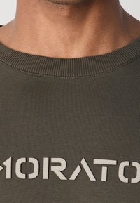 Olivgrön tygtröja med ribbad halsringning och texturerad vit logotypdetalj över framsidan. Fokusera på den släta, mjuka texturen.