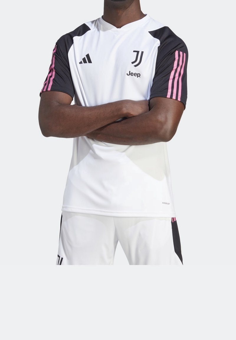 Personne portant un maillot de football blanc de la Juventus avec des manches noires et roses, bras croisés, assorti à un short blanc.