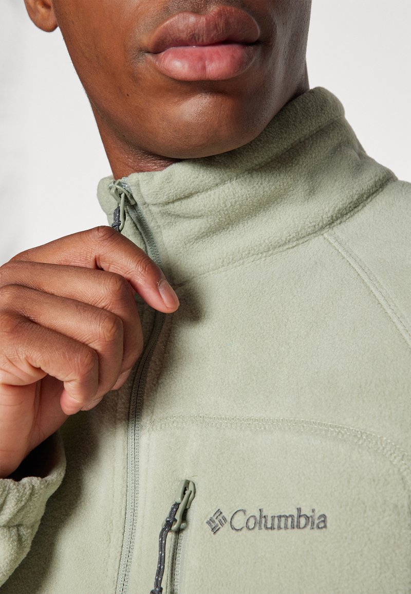 Chaqueta de forro polar verde con un cuello alto, cierre frontal con cremallera y logotipo de Columbia en el pecho. Tela texturizada y tirador de cremallera ajustable.