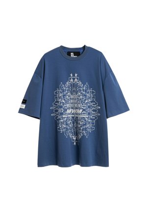 Persona con rastas que lleva una camisa azul de gran tamaño con un gran gráfico de una mujer rezando con ojos azules y tatuajes en la espalda, pantalones negros y zapatos.