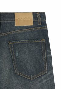 Donkerdenim jeans met een lichtbruine leren label met de tekst "HUGO." Voorzien van oranje stiksels en een enkele achterzak met beschadigde details.