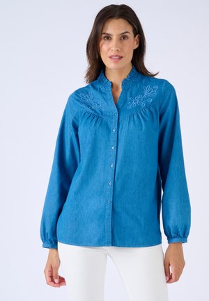 Femme portant un chemisier bleu à manches longues boutonné avec un col à volants et une broderie florale blanche sur la poitrine, associé à un pantalon blanc.