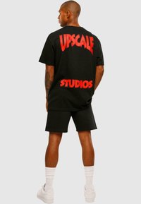 Čierne oversized tričko so výrazným červeným textom "UPSCALE" a "STUDIOS" na chrbte, v kombinácii s čiernymi šortkami a bielymi teniskami.