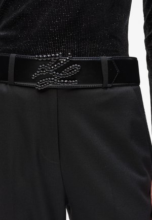 Schwarzer Gürtel mit glänzender Oberfläche, der ein mit Perlen gestaltetes Muster aufweist, das einer Schlange ähnelt. Wird über dunklem Stoff getragen und betont die Textur und Details.