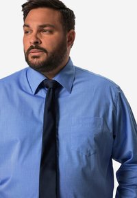 Světle modrá košile na knoflíky s kapsou, doplněná tmavomodrou kravatou. Materiál vypadá hladce s jemnou strukturou.