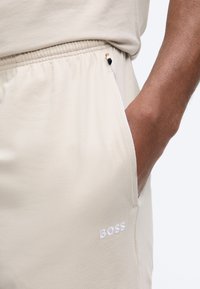 Μπεζ παντελόνι φόρμας με ελαστική μέση και πλαϊνή τσέπη, χέρι στην τσέπη, και κεντημένο λογότυπο "BOSS" κοντά στην τσέπη.