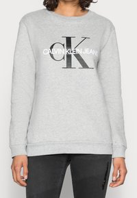 Felpa grigia con colletto a giro, caratterizzata da un grande logo nero che riporta la scritta "CALVIN KLEIN JEANS" in stampa grassetto sulla parte anteriore.