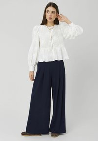 Blouse blanche texturée avec manches bouffantes et ourlet volanté, associée à un pantalon marine taille haute à jambes larges. Le mannequin pose avec une main levée.