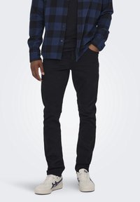 Jeans neri con una vestibilità slim, abbinati a una camicia a quadri blu con bottoni. Sneakers bianche con dettagli blu navy completano l'outfit.