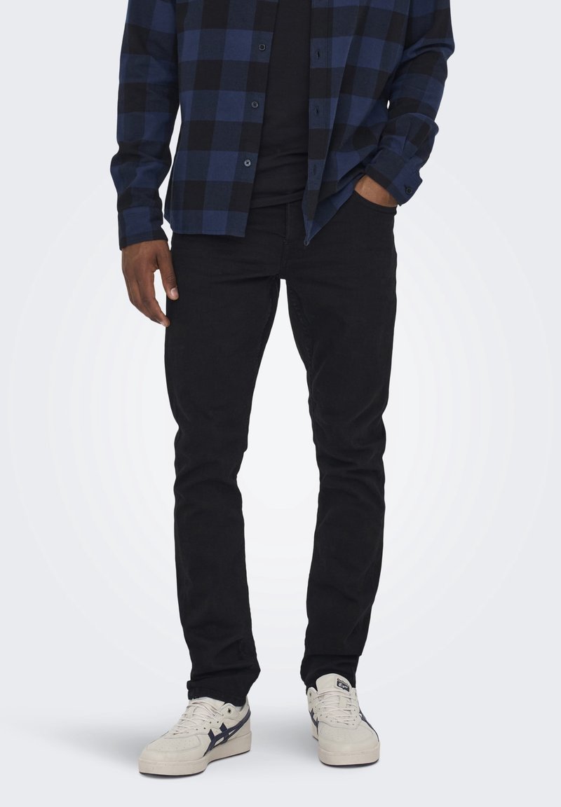 Jeans neri con una vestibilità slim, abbinati a una camicia a quadri blu con bottoni. Sneakers bianche con dettagli blu navy completano l'outfit.