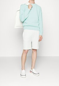 Minzgrüner, strukturiert Pullover, helle Shorts und weiße Slipper mit roten Akzenten. Der Hintergrund ist weiß.