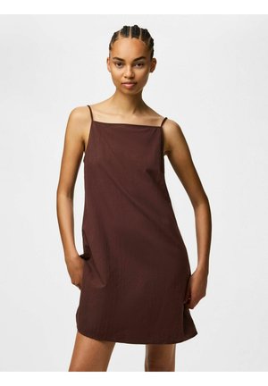 Jeune femme aux cheveux tressés portant une mini-robe marron foncé sans manches, debout les mains dans les poches devant un fond uni.