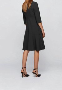 Svart figurnära klänning med trekvartsärmar, utsvängd fåll och detaljer på bakstycket. Bärs med svarta stilettklackar. Slät tygtextur.