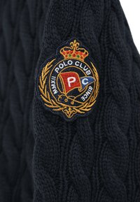 Stickad marinblå tröja med ett texturerat kabelmönster. Inkluderar en guld- och färgbroderad badge med en krona och texten "Polo Club."