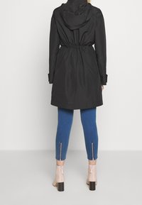 Impermeabile nero con cappuccio e coulisse in vita, abbinato a jeans skinny blu con cerniere alle caviglie e stivaletti beige con tacco.