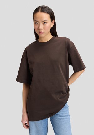 Femme aux longs cheveux foncés portant une chemise ample marron foncé à manches courtes et un jean bleu clair, debout devant un fond clair uni.