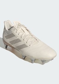 Vita fotbollssko med strukturerad ovandel, tonala tre ränder-logotyp, snörning och genomskinliga dobbar på en grå sula.