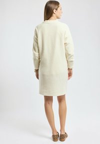 Robe en tricot couleur crème avec des manches longues, un col rond et un motif texturé. Portée avec des chaussures à imprimé animal marron.