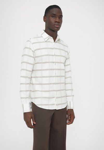 Wit overhemd met knopen, grijs ruitpatroon, zachte stof, lange mouwen en een afgeronde zoom. Model draagt een bruine broek.