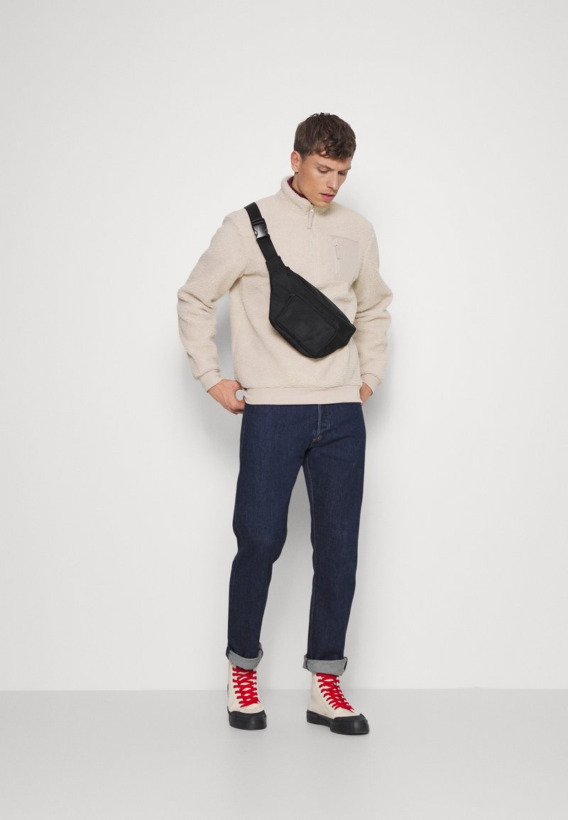Schwarze Umhängetasche aus strapazierfähigem Material, mit einem Reißverschlussfach und verstellbarem Gürtel, getragen über einem beigen Fleece-Pullover und blauen Jeans.