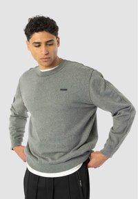 Hombre ajustando un suéter gris de cuello redondo sobre una camisa blanca, vistiendo pantalones negros con cremallera lateral, mirando hacia abajo y ligeramente hacia la izquierda.