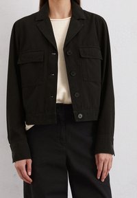 Svart kort jacka i bomull med krage, knappstängning fram och två bröstfickor. Strukturerad tyg med en avslappnad design.