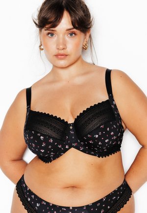 Femme à la peau claire et aux yeux bleus porte un soutien-gorge en dentelle noire à motifs floraux et des sous-vêtements assortis, debout devant un fond blanc.
