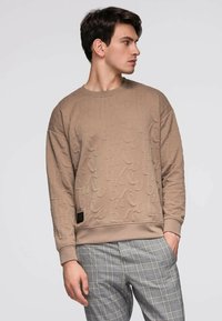 Beige collegepaita kohokuvioiduilla kuvioilla, pyöreällä pääntiellä ja resorilla varustetuilla hihoilla. Yhdistetty harmaisiin ruutukuviollisiin housuihin, jotka korostavat tekstuurin kontrastia.