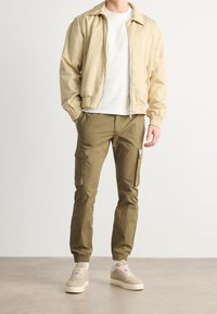 Beige lättviktsjacka med elastiska mudd, bärs över en vit skjorta, tillsammans med olivgröna cargobyxor med sidofickor och beige sneakers.