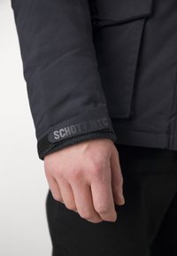 Gros plan sur la main d'une personne portant une veste noire avec un bracelet Velcro étiqueté "SCHOTT NYC" sur une manche intérieure noire.
