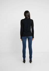 Suéter de cuello alto de manga larga negro, diseño ajustado, textura suave, combinado con jeans ajustados azules y botas negras hasta el tobillo. Vista trasera.