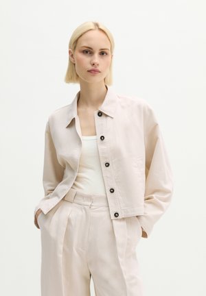 Femme aux cheveux blonds courts portant une veste boutonnée beige clair sur un haut blanc et un pantalon taille haute assorti, les mains dans les poches.