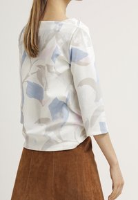 Witte blouse met bloemige patronen in blauwe en roze tinten, driekwart mouwen, textiel stof, gedragen met een bruine suède rok.