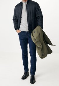 Svart vadderad bomberjacka över en grå tröja, kombinerad med mörkblå jeans och svarta läderstövlar. Olivgrön kappa hålls i handen.