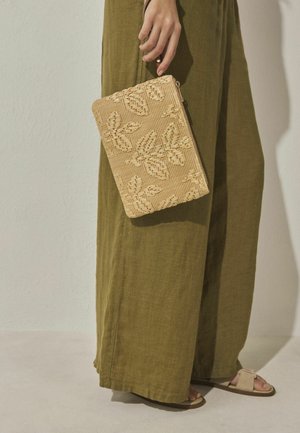 Hand holding beige woven clutch with butterfly patterns, wearing olive-green wide-leg pants and beige sandals on light floor.