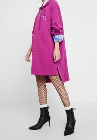 Person som bär en ljuslila oversized huvtröja i klänningstil, nätstrumpbyxor, vita strumpor och svarta spetiga högklackade ankelstövlar.