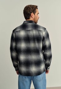 Camicia a scacchi nera e grigia con una texture morbida, polsini con bottone e orlo inferiore curvo, indossata su jeans azzurri chiari.